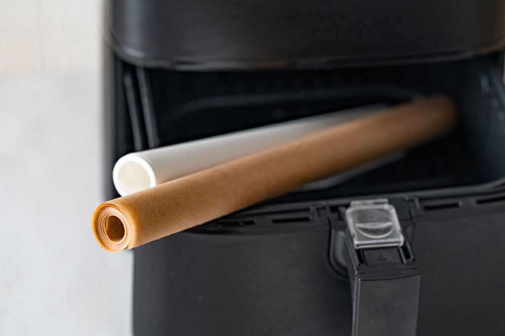 Zwei Backpapier-Rollen liegen im halb geöffneten Korb einer Heißluftfritteuse bzw. Airfryer.